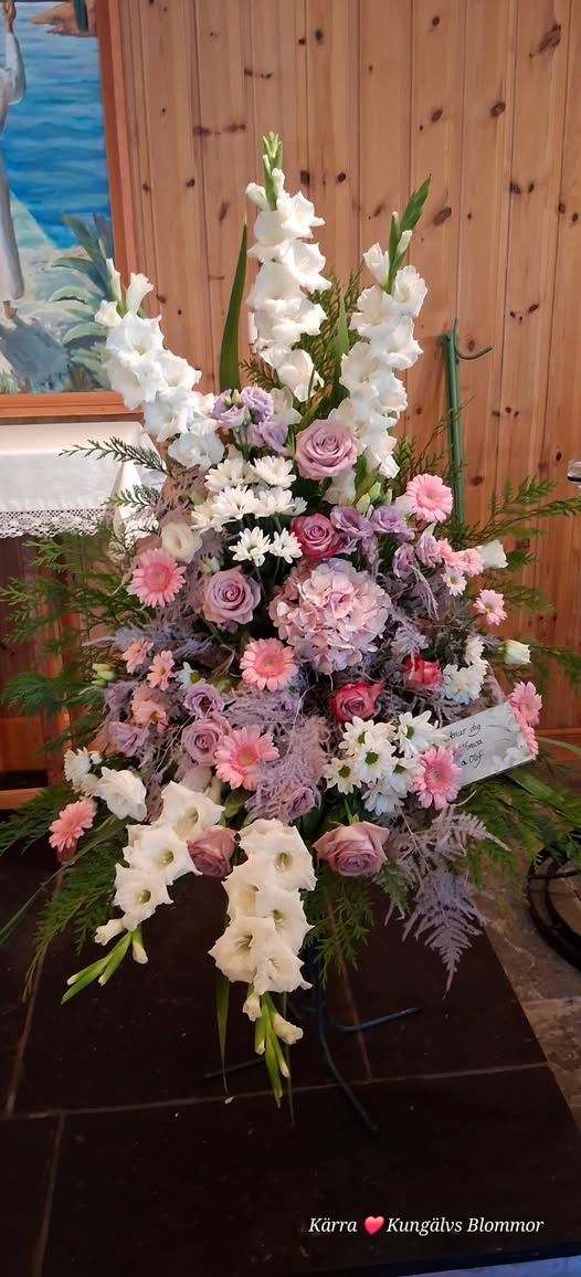 Ett stort begravningsbinderi med vita gladiolus, lila rosor, rosa gerbera och ormbunke framför en träpanel